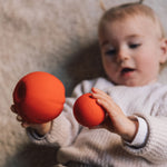 A Baby holding onto two Oli & Carol Tomato balls, one large one small. 