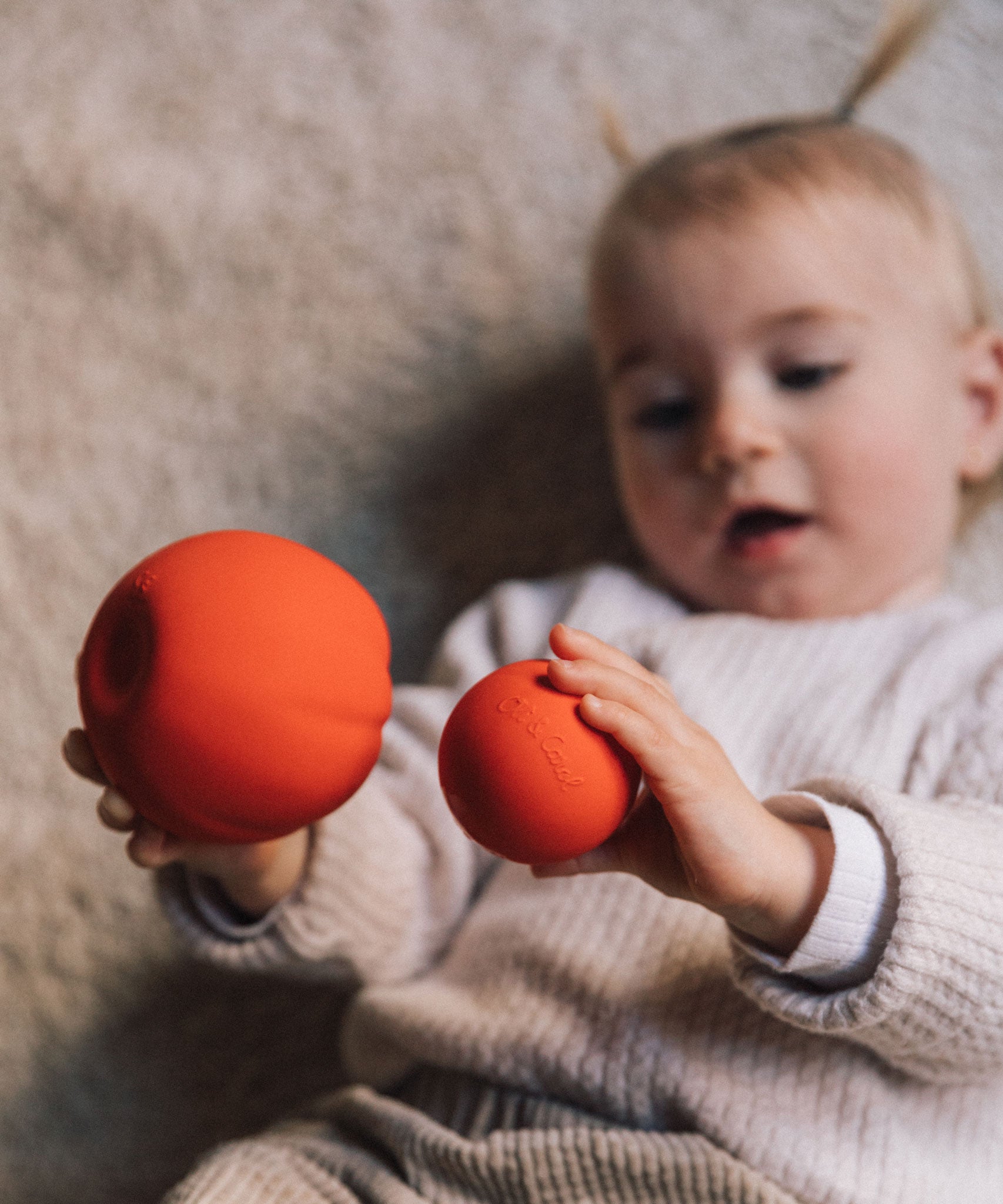 A Baby holding onto two Oli & Carol Tomato balls, one large one small. 