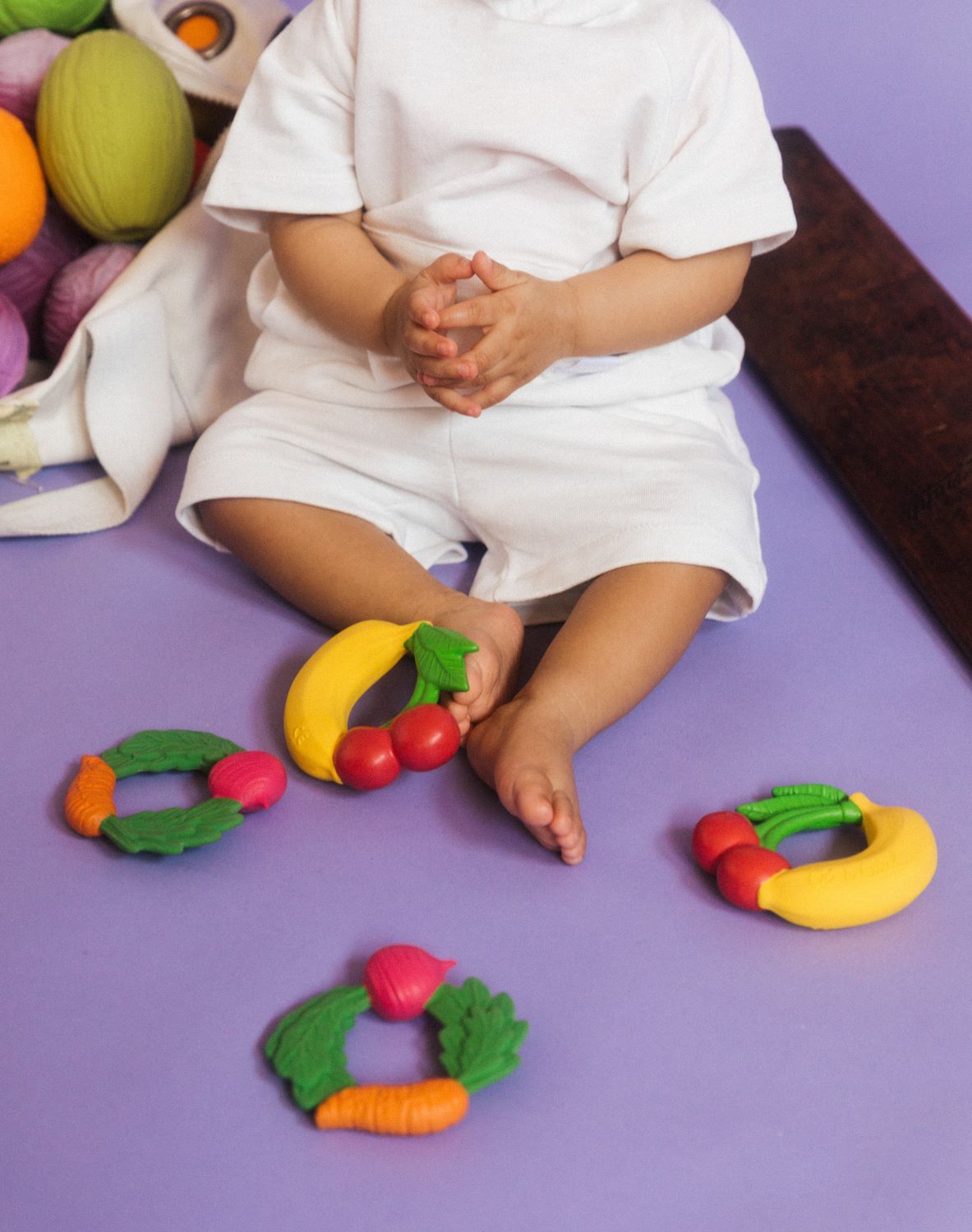 4 Oli & Carol Natural Rubber Veggie and fruit Teething Rings on the floor, a child can be seen sitting in the background. 