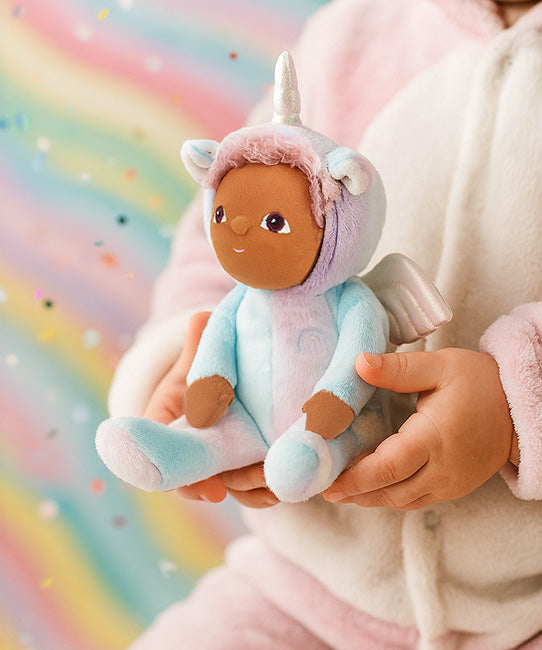 Olli Ella Dinky dinkum doll Uli Unicorn available at Babipur. Close up of soft cuddly doll held by a child with a glittery rainbow background.  