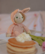 Olli Ella pink coloured Babbit Bunny Dinky Dinkum Doll in a seated position on a blanket. A pile of pancakes can be seen at the front. 