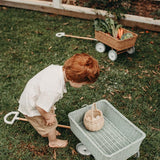 Boy hunched over collecting eggs in a basket inside the Olli Ella blue rattan wagon on some grass