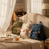 Olli Ella cozy bag charms clipped on 3 backpacks sitting on top of a bed showing scale. These bag charms are available at Babipur.