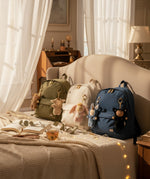 Olli Ella cozy bag charms clipped on 3 backpacks sitting on top of a bed showing scale. These bag charms are available at Babipur.