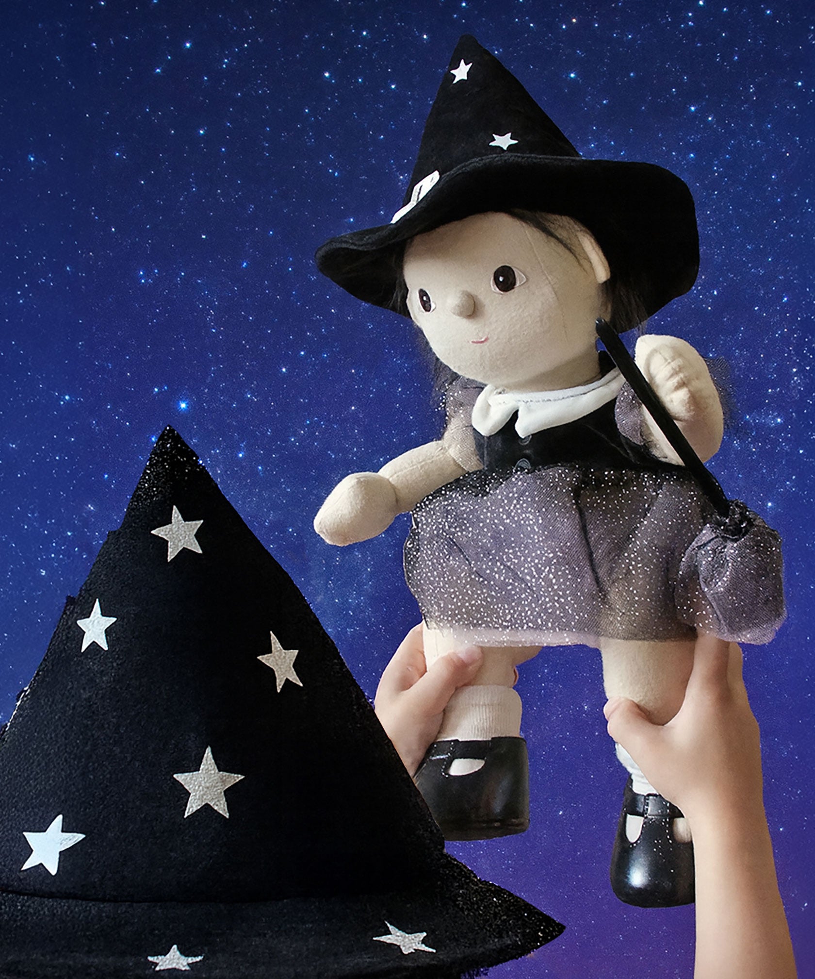 A child holding up an Olli Ella Wendy Witch dream Dinkum doll. A dinkum doll with a black dress, witches hat, black shoes and a magnetic broom accessory. This doll is part of Olli Ella's Halloween collection available here at Babipur.