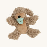 A soft and fluffy Brown Olli Ella Benny puppy with a magnetic dog toy in its mouth on a cream background.