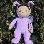 The Olli Ella Dinky Dinkum Doll - Betty Butterfly - in a light purple onesie, with brown hair, blue eyes, dark purple butterfly wings, and antennae. She is in a green leafy bush.