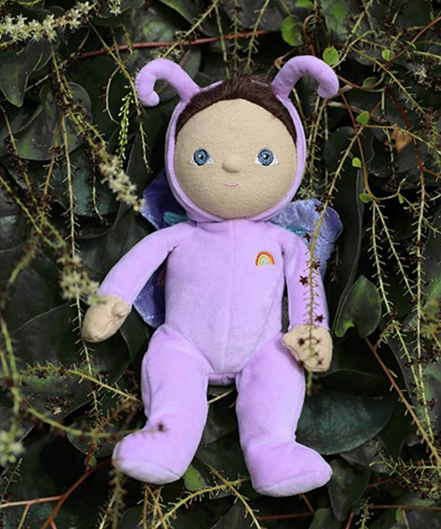 The Olli Ella Dinky Dinkum Doll - Betty Butterfly - in a light purple onesie, with brown hair, blue eyes, dark purple butterfly wings, and antennae. She is in a green leafy bush.