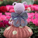 The Olli Ella Dinky Dinkum Doll - Betty Butterfly - in a light purple onesie, with brown hair, blue eyes, dark purple butterfly wings, and antennae. It is sat on a rattan toadstool basket in a flower garden.