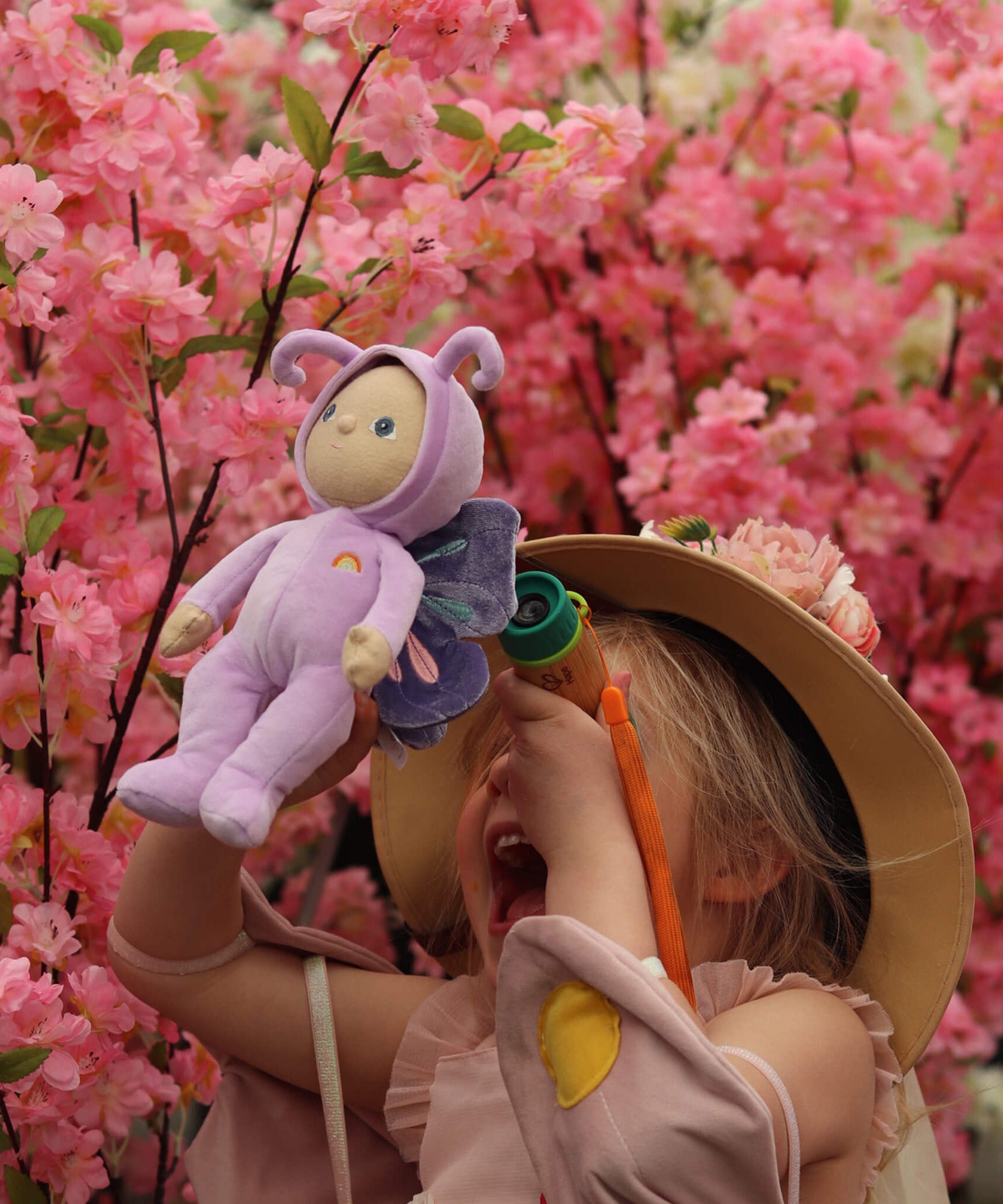 A little girl holding the Olli Ella Dinky Dinkum Doll - Betty Butterfly -in a pink flower garden.
