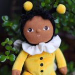 The Olli Ella Dinky Dinkum Doll - Billie Bumblebee - in a yellow onesie with a black hood and antennae, with a white ruffled collar, brown eyes, curly black hair, and buzzy bee wings.