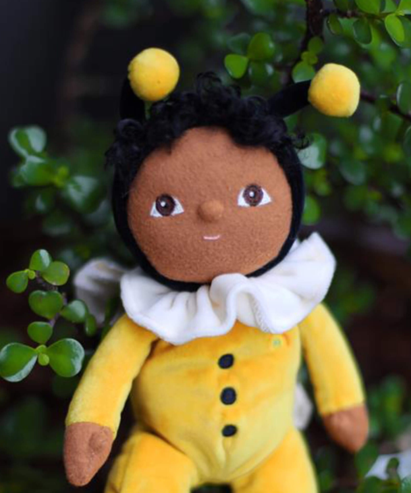 The Olli Ella Dinky Dinkum Doll - Billie Bumblebee - in a yellow onesie with a black hood and antennae, with a white ruffled collar, brown eyes, curly black hair, and buzzy bee wings.