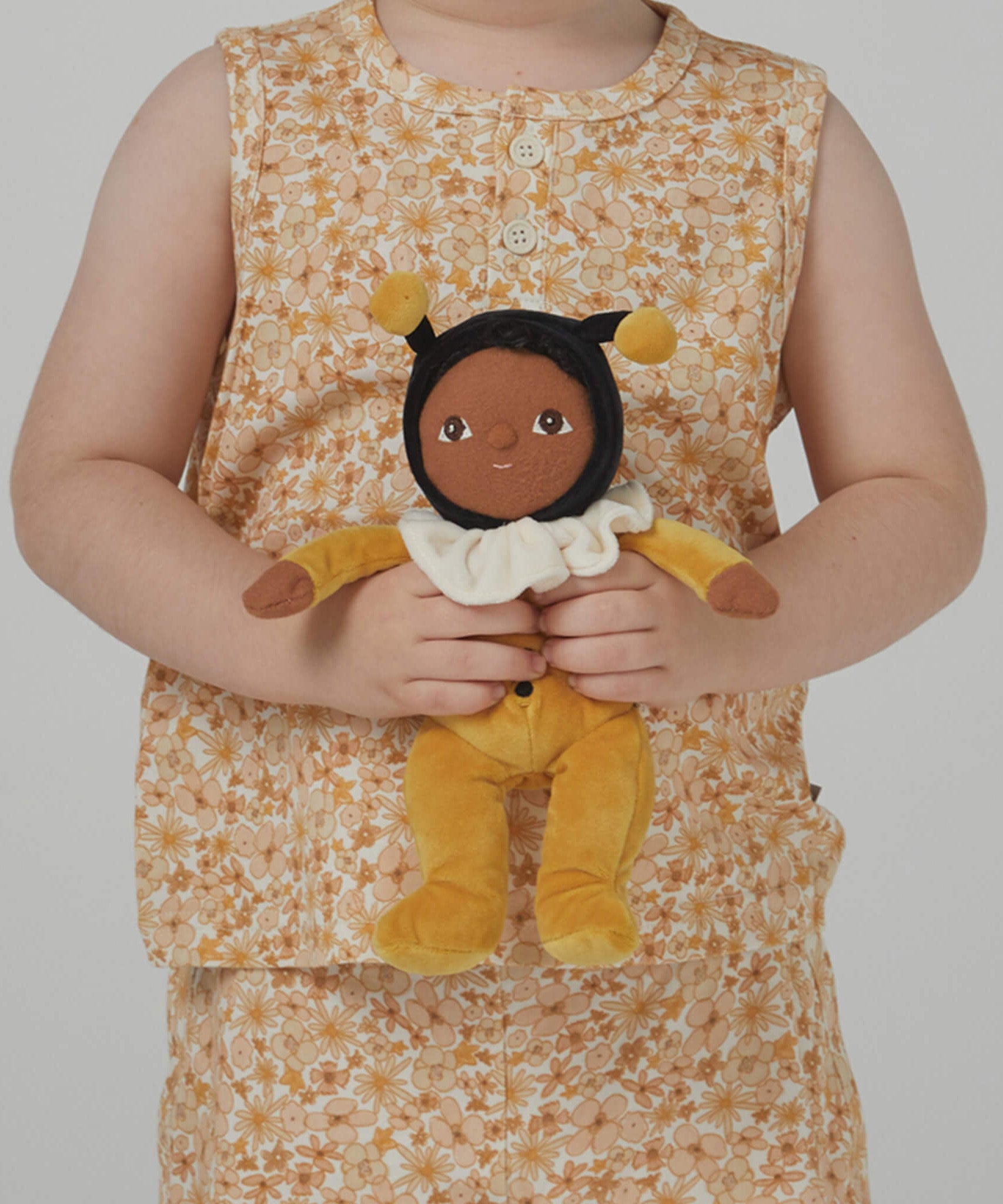 A little girl holding the Olli Ella Dinky Dinkum Doll - Billie Bumblebee - in a yellow onesie with a black hood and antennae, with a white ruffled collar, brown eyes, and buzzy bee wings.