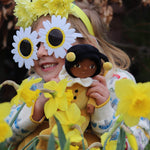 A little girl in flower sunglasses, holding the Olli Ella Dinky Dinkum Doll - Billie Bumblebee - in a yellow onesie with a black hood and antennae.