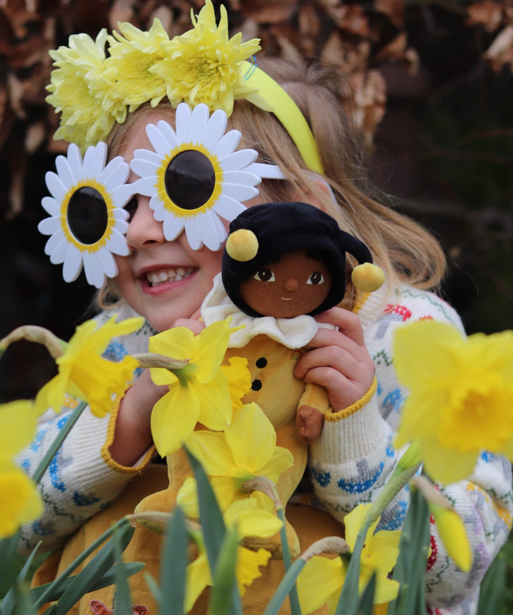A little girl in flower sunglasses, holding the Olli Ella Dinky Dinkum Doll - Billie Bumblebee - in a yellow onesie with a black hood and antennae.