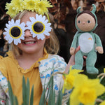 A little girl in flower sunglasses holding the Olli Ella Dinky Dinkum Doll - Gideon Grasshopper - in a garden.
