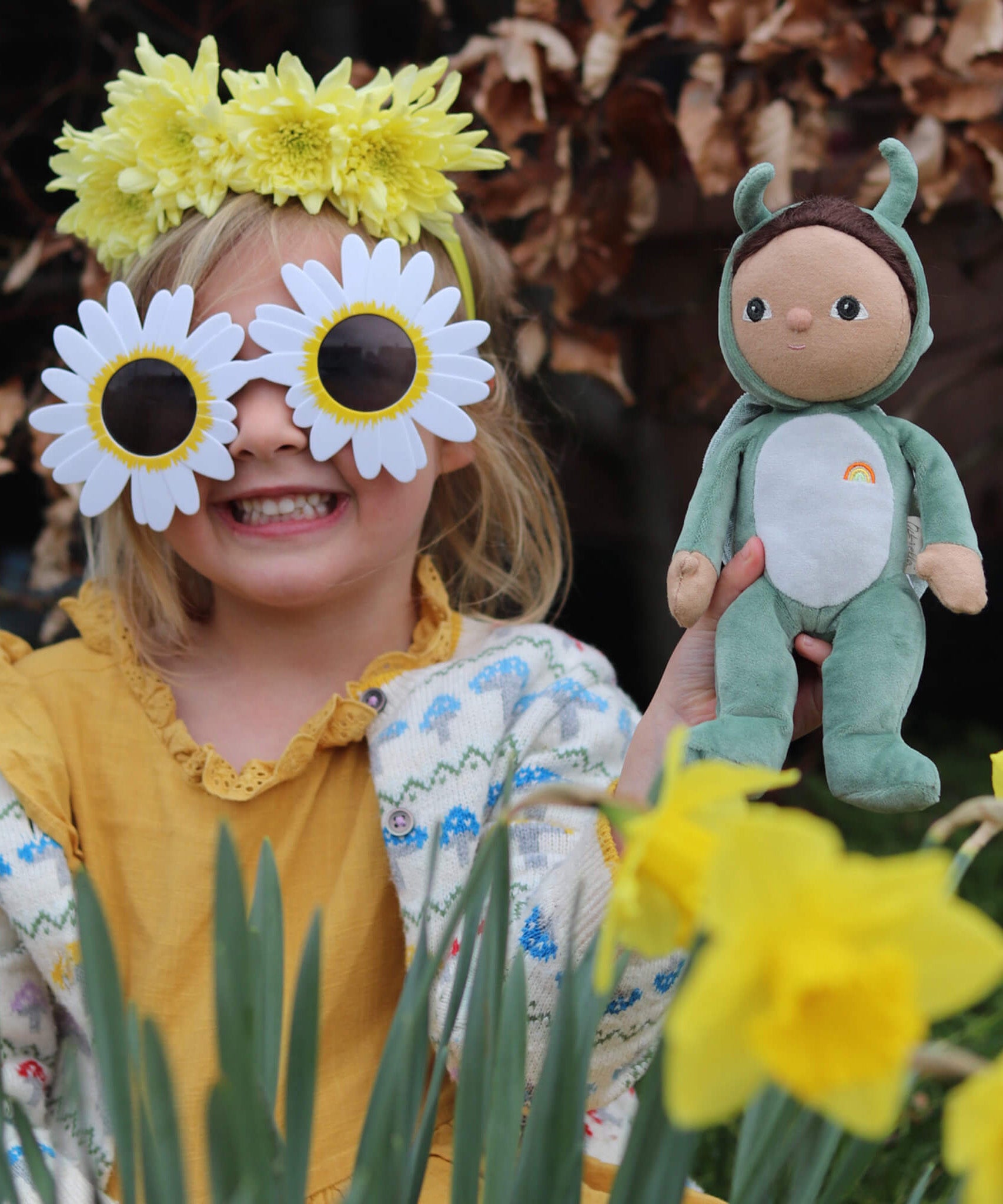 A little girl in flower sunglasses holding the Olli Ella Dinky Dinkum Doll - Gideon Grasshopper - in a garden.