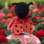 The back of the Olli Ella Dinky Dinkum Doll - Lola Ladybug - in a red onesie with ladybird wings, sat on a rattan toadstool basket in a flower garden.