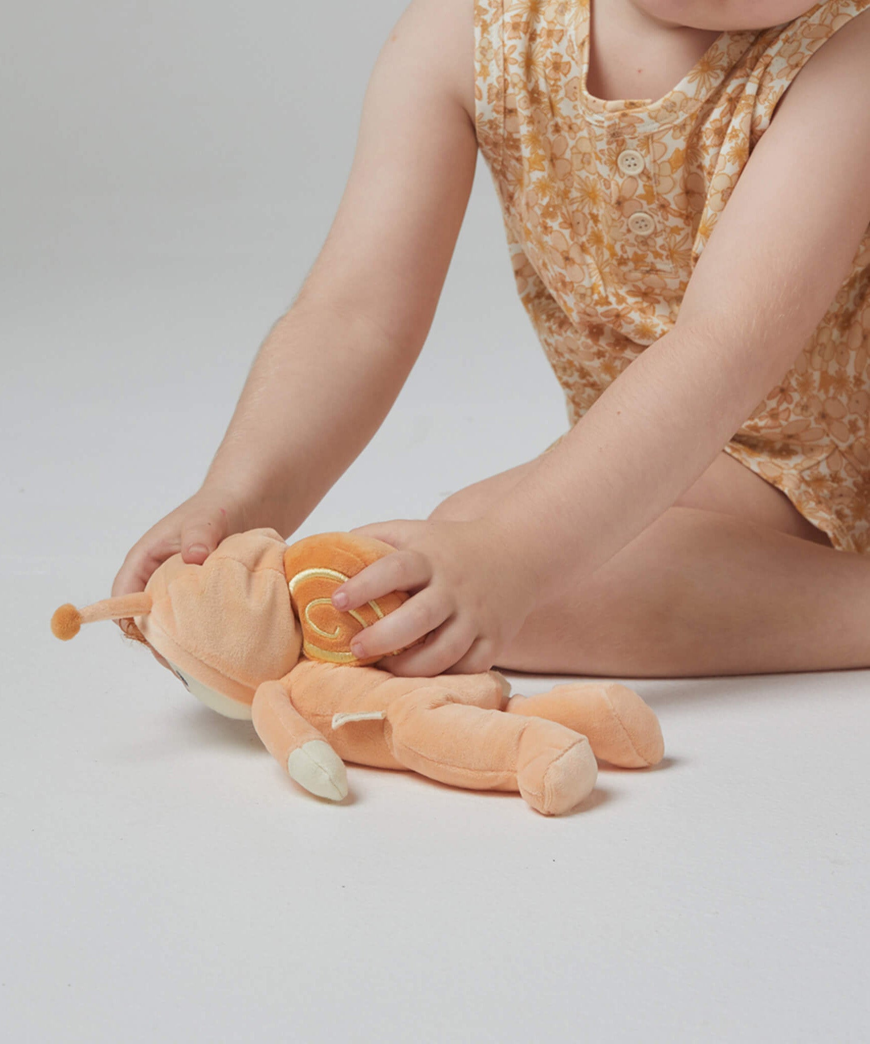 A little girl playing with the Olli Ella Dinky Dinkum Doll - Scottie Snail - in a soft light orange onesie with a snail shell on his back.