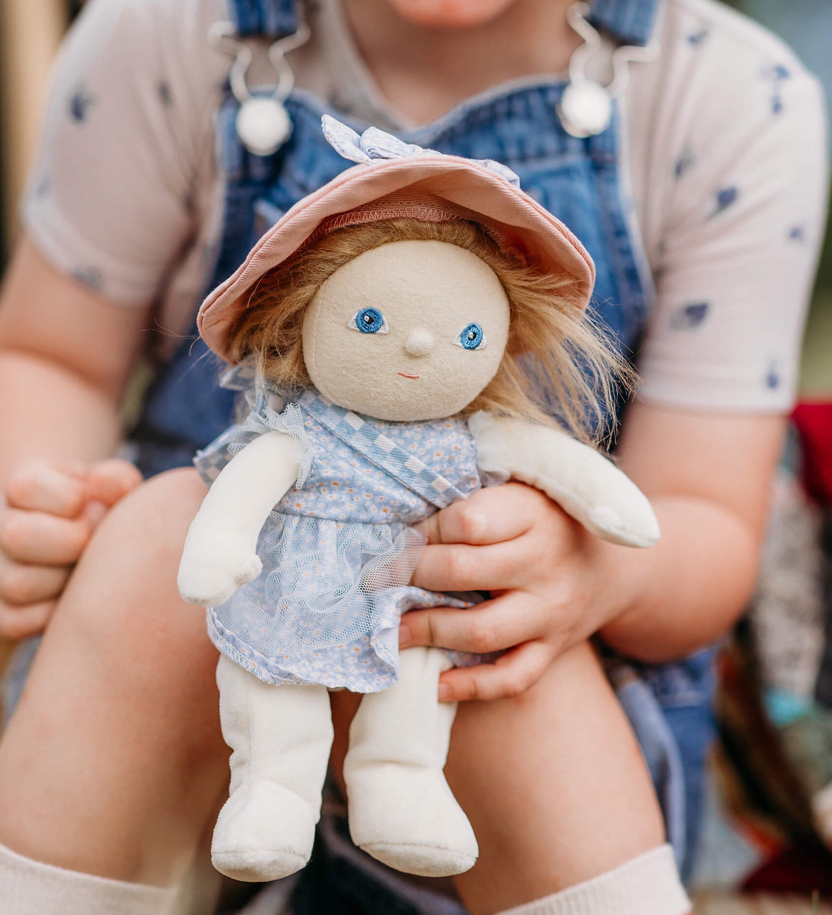 Olli Ella Dress Me Dinky Dinkum Doll Penelope Petals held by a child, showing her blue floral dress, pink sunhat, and soft posable design.
