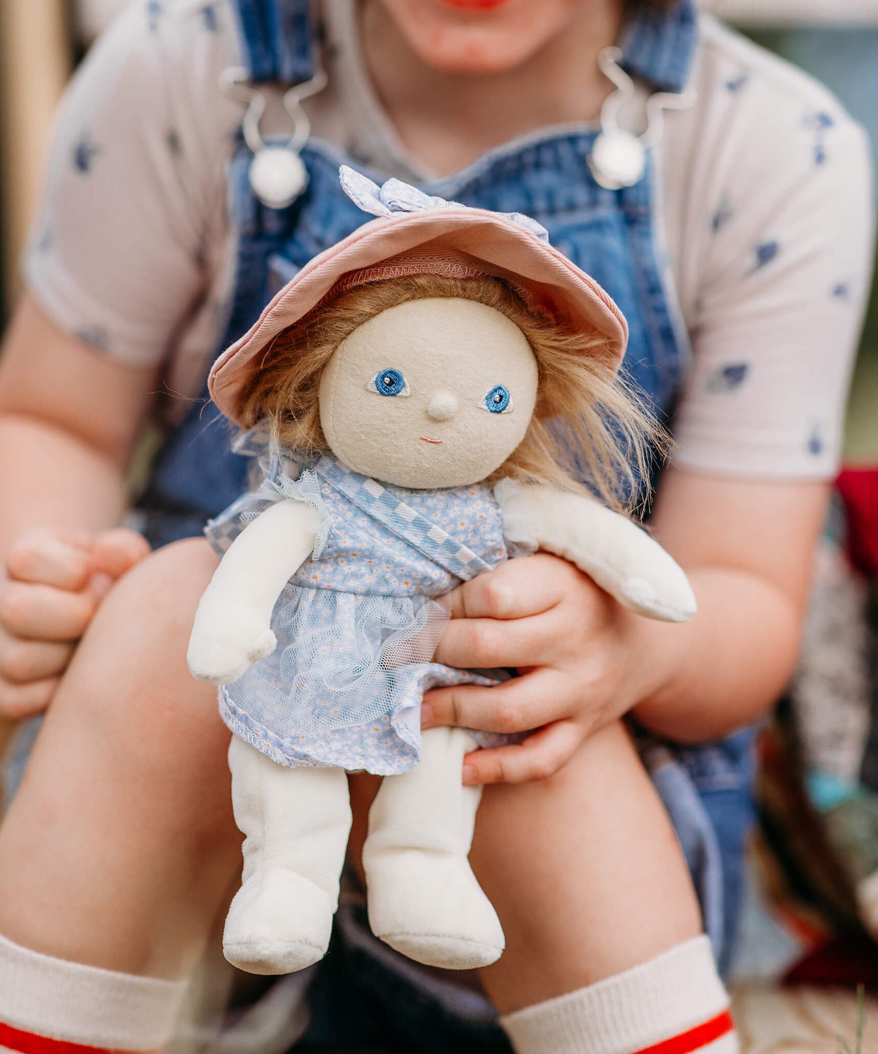 Olli Ella Dress Me Dinky Dinkum Doll Penelope Petals held by a child, showing her blue floral dress, pink sunhat, and soft posable design.