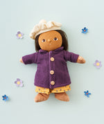 Olli Ella Dress Me Dinky - Lila Lavender wearing a purple fleece trench coat, yellow dress and white beret. Surrounded by paper flowers, blue background. 