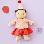 Olli Ella Dress Me Dinky - Sophie Sundae, a doll with brown hair and green eyes wearing a pink polka dot dress, light pink cardigan and a pink party hat. Holding a paper balloon and present, purple background.