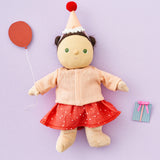 Olli Ella Dress Me Dinky - Sophie Sundae, a doll with brown hair and green eyes wearing a pink polka dot dress, light pink cardigan and a pink party hat. Holding a paper balloon and present, purple background.