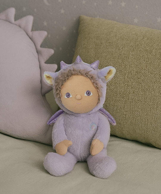 olli ella duffy the dinky dragon soft cuddly doll available at babipur. doll is sitting on a blanket with cushions in the background.