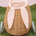 A child happily stood in the sun next to the Olli Ella Rattan Bunny Luggy with Lining – Gumdrop and holding the Rattan basket in Gumdrop