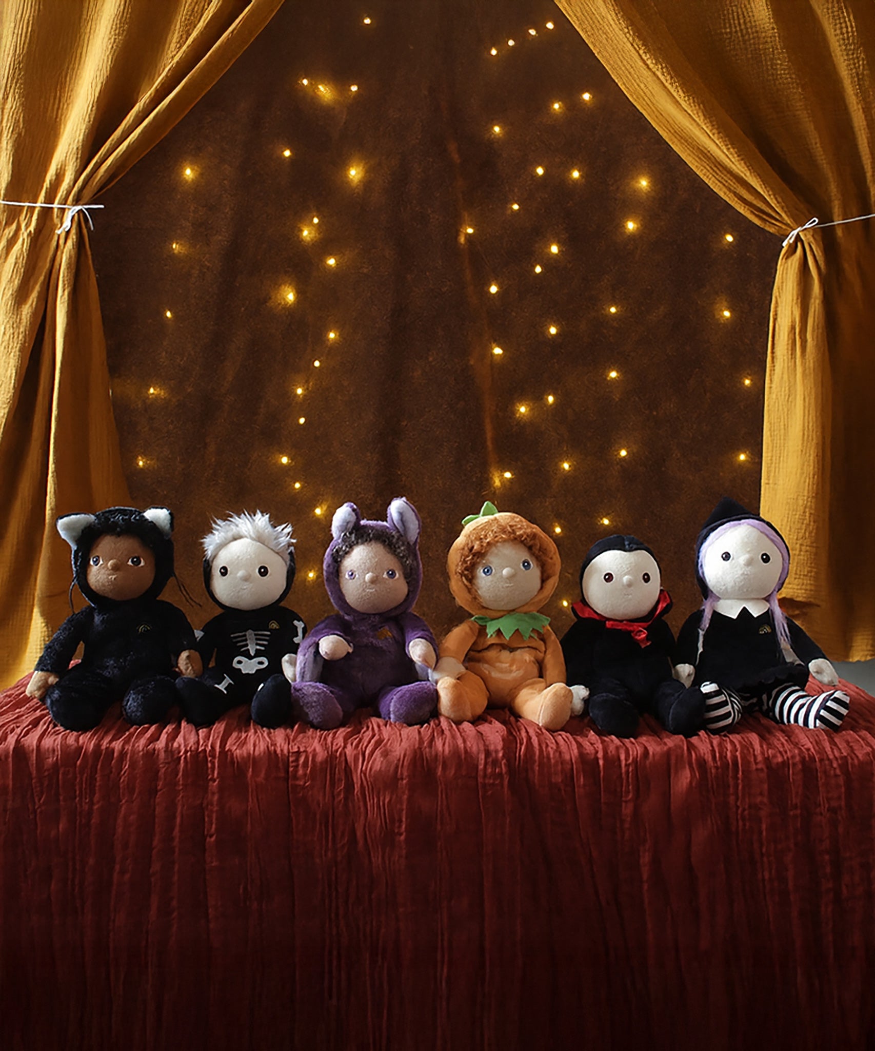A collection of six Halloween themed Olli Ella dinky dinkums all lined up on a surface covered with a dark red coloured cloth. These soft toys are part of Olli Ella's Halloween collection available here at Babipur.