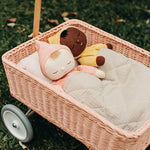 Close up of 2 Olli Ella dinkum dolls laying in the rose pink rattan toy wagon on some grass