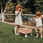 Girl and boy stood on some grass next to the Olli Ella light pink rattan wonder wagon toy
