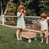 Girl and boy stood on some grass next to the Olli Ella light pink rattan wonder wagon toy