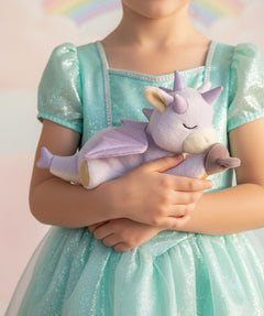 A child wearing a princess outfit and holding a Olli Ella misty binky dragon doll. This light purple fluffy dragon has a sleeping face and features glittery wings and comes with a purple magnetic pacifier. This soft toy is available at Babipur.