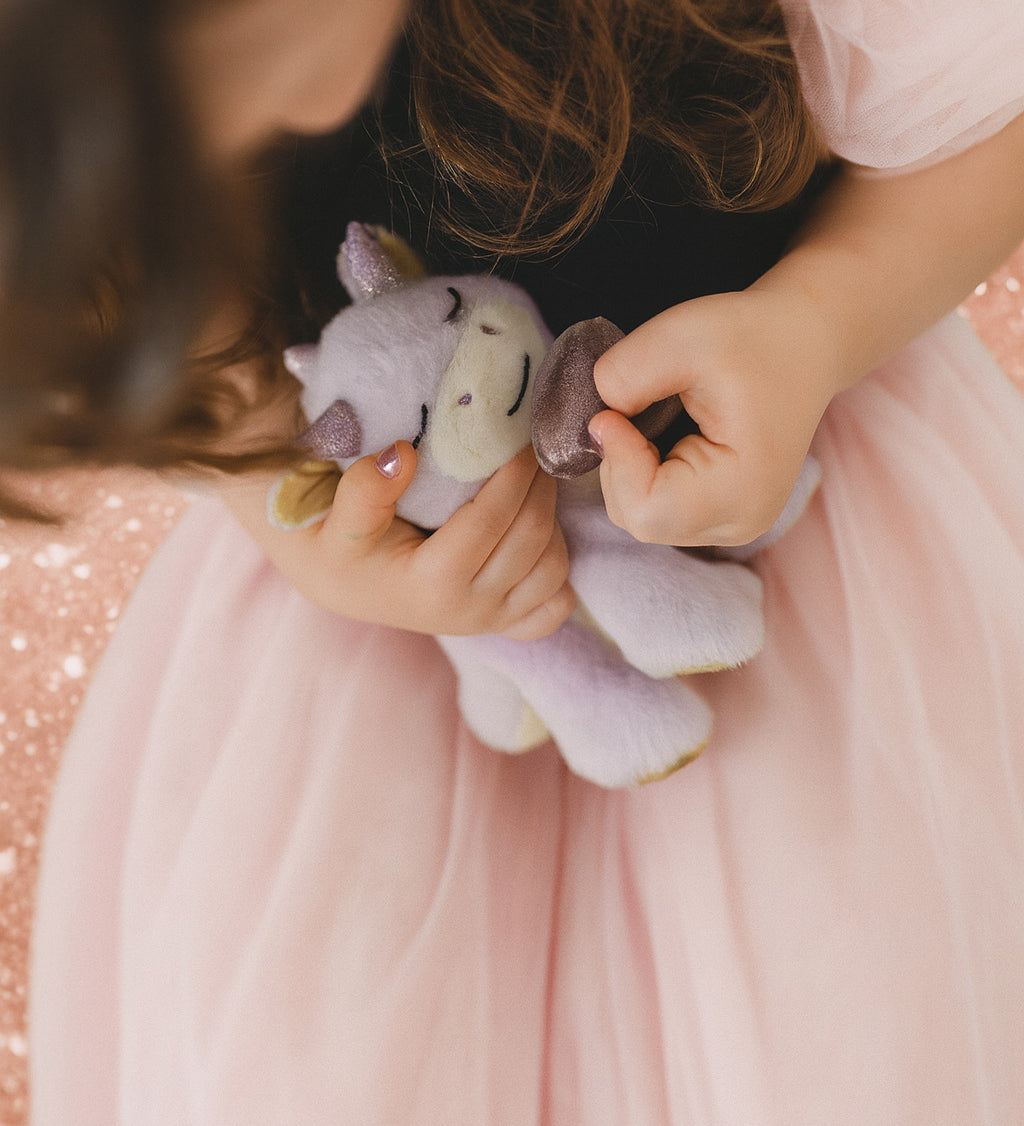 A child wearing a princess outfit and holding a Olli Ella misty binky dragon doll with the magnetic purple pacifier near the mouth. This fluffy soft toy is available at Babipur.