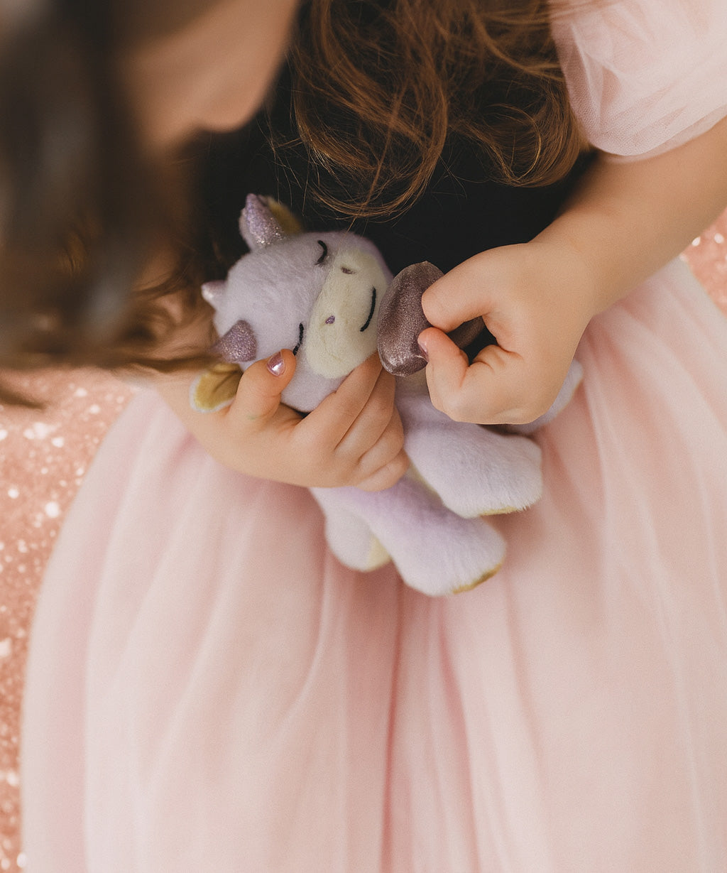 A child wearing a princess outfit and holding a Olli Ella misty binky dragon doll with the magnetic purple pacifier near the mouth. This fluffy soft toy is available at Babipur.