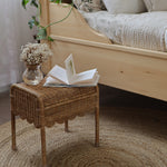 Olli Ella Petal Storie Stool in Natural Rattan. The stool is shown next to a wooden bed with an open book and small glass vase of flowers besides it. 
