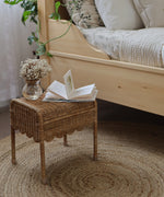 Olli Ella Petal Storie Stool in Natural Rattan. The stool is shown next to a wooden bed with an open book and small glass vase of flowers besides it. 