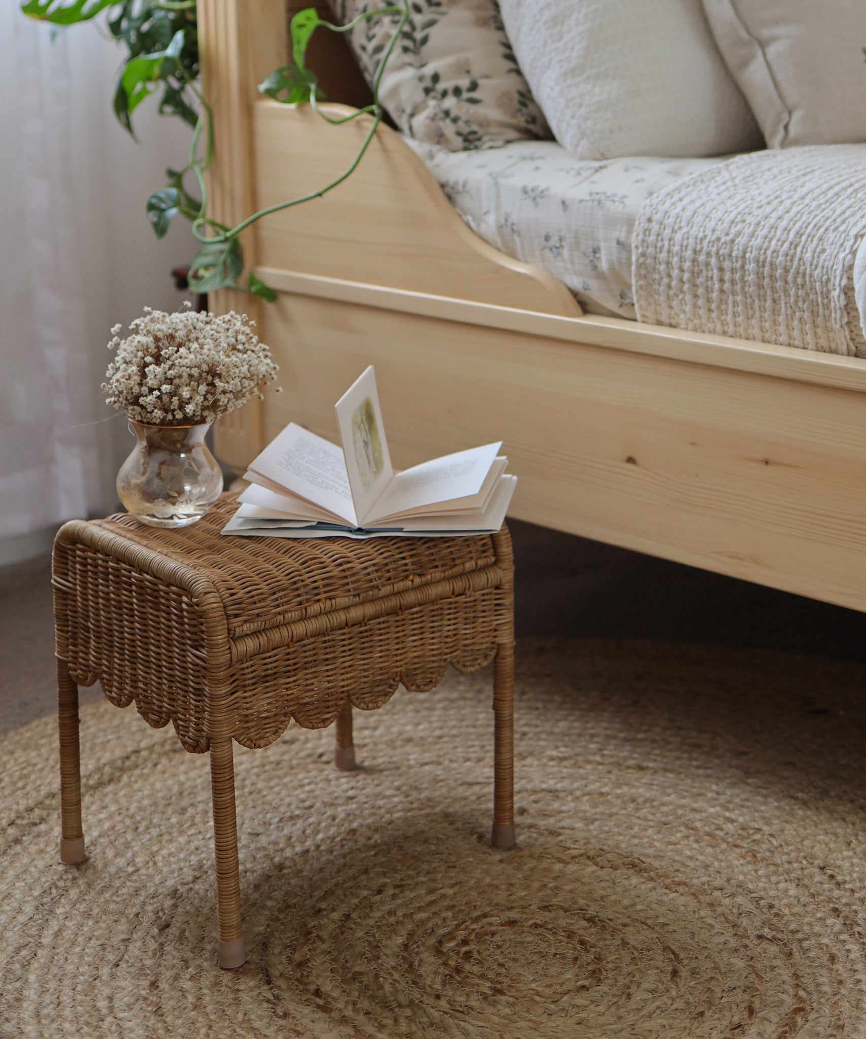 Olli Ella Petal Storie Stool in Natural Rattan. The stool is shown next to a wooden bed with an open book and small glass vase of flowers besides it. 