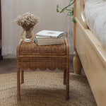 Olli Ella Petal Storie Stool in Natural Rattan. The stool has been placed by the side of a wooden bed and has books piled on the top. 