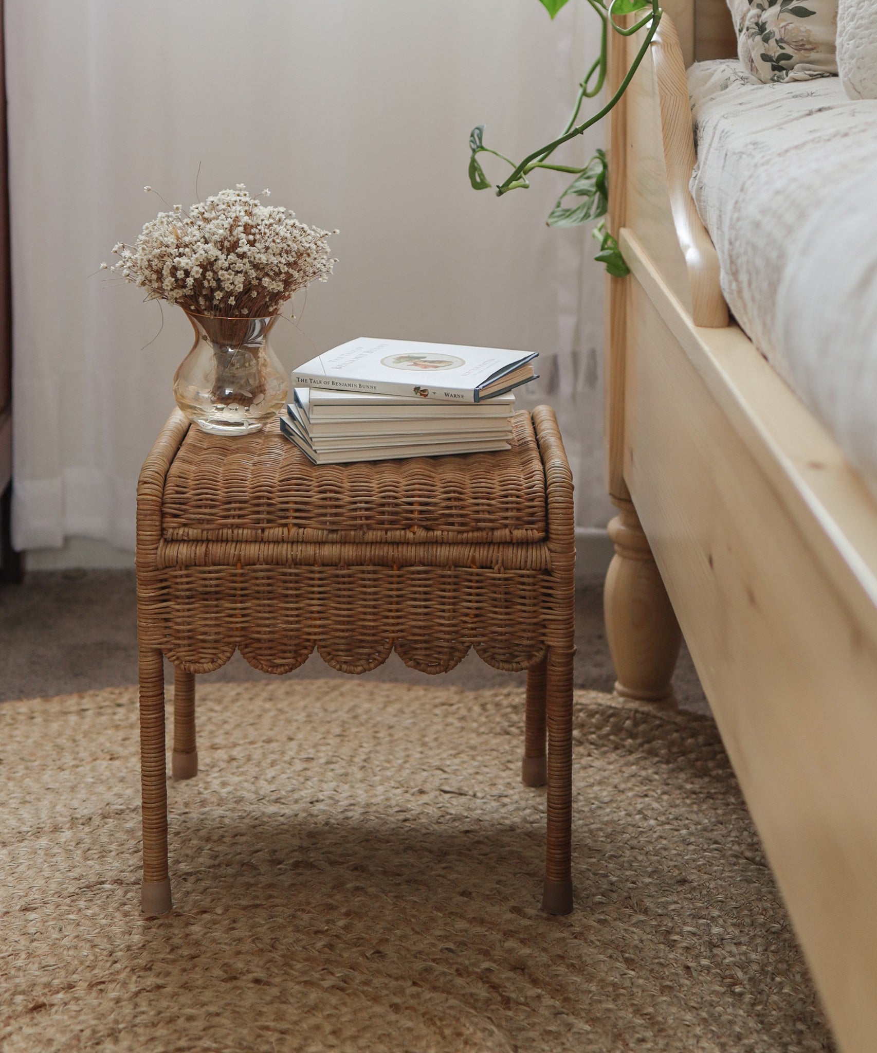 Olli Ella Petal Storie Stool in Natural Rattan. The stool has been placed by the side of a wooden bed and has books piled on the top. 