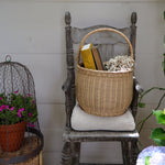 An Olli Ella Large Natural Rattan Gathering basket placed on a chair. There are books and flowers in the basket. 