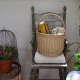 An Olli Ella Large Natural Rattan Gathering basket placed on a chair. There are books and flowers in the basket. 