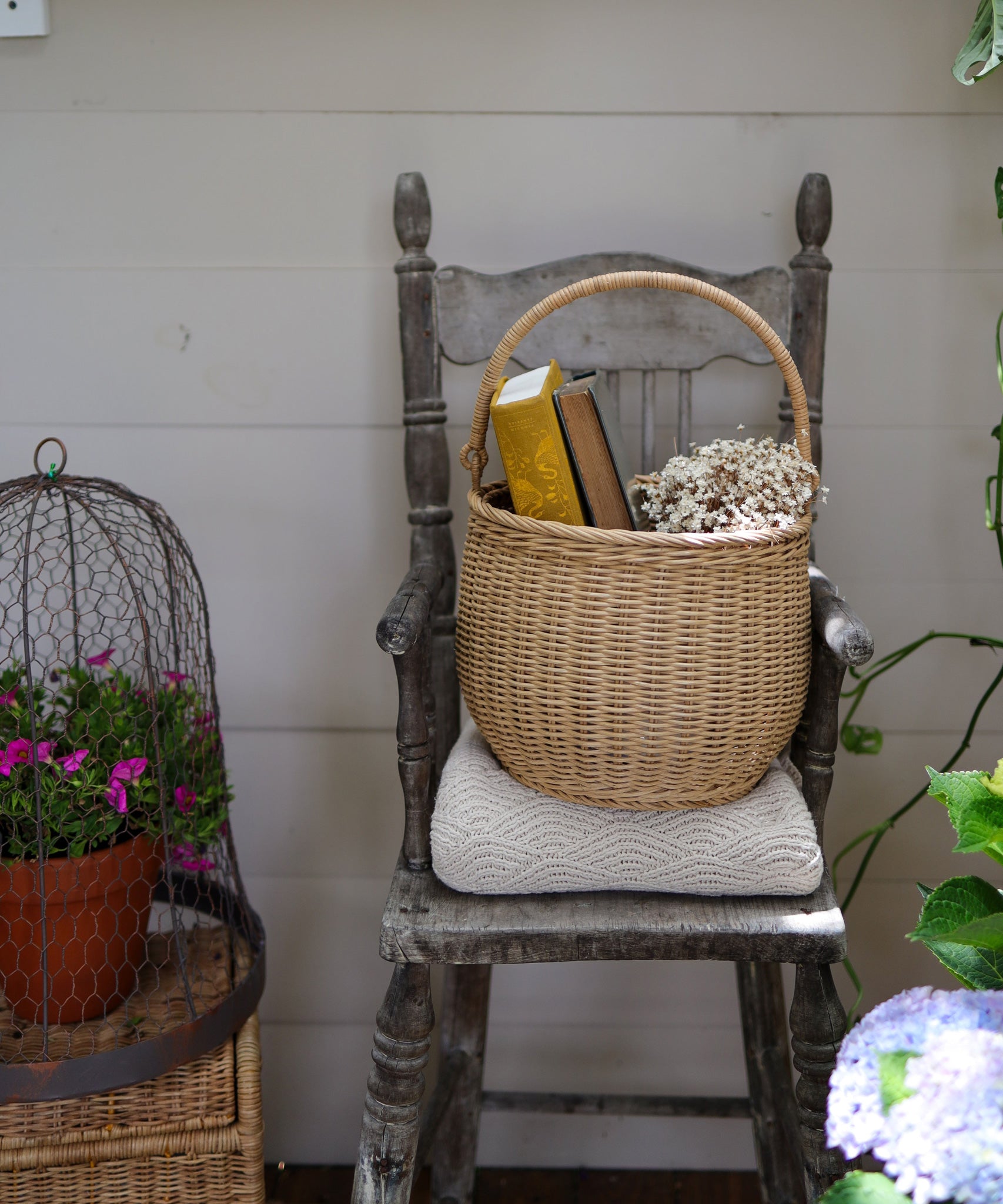 An Olli Ella Large Natural Rattan Gathering basket placed on a chair. There are books and flowers in the basket. 