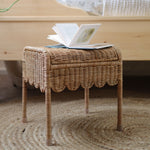 A Olli Ella Petal Storie Stool in natural Rattan. The stool has been placed by the side of a bed with an open book placed on top. 
