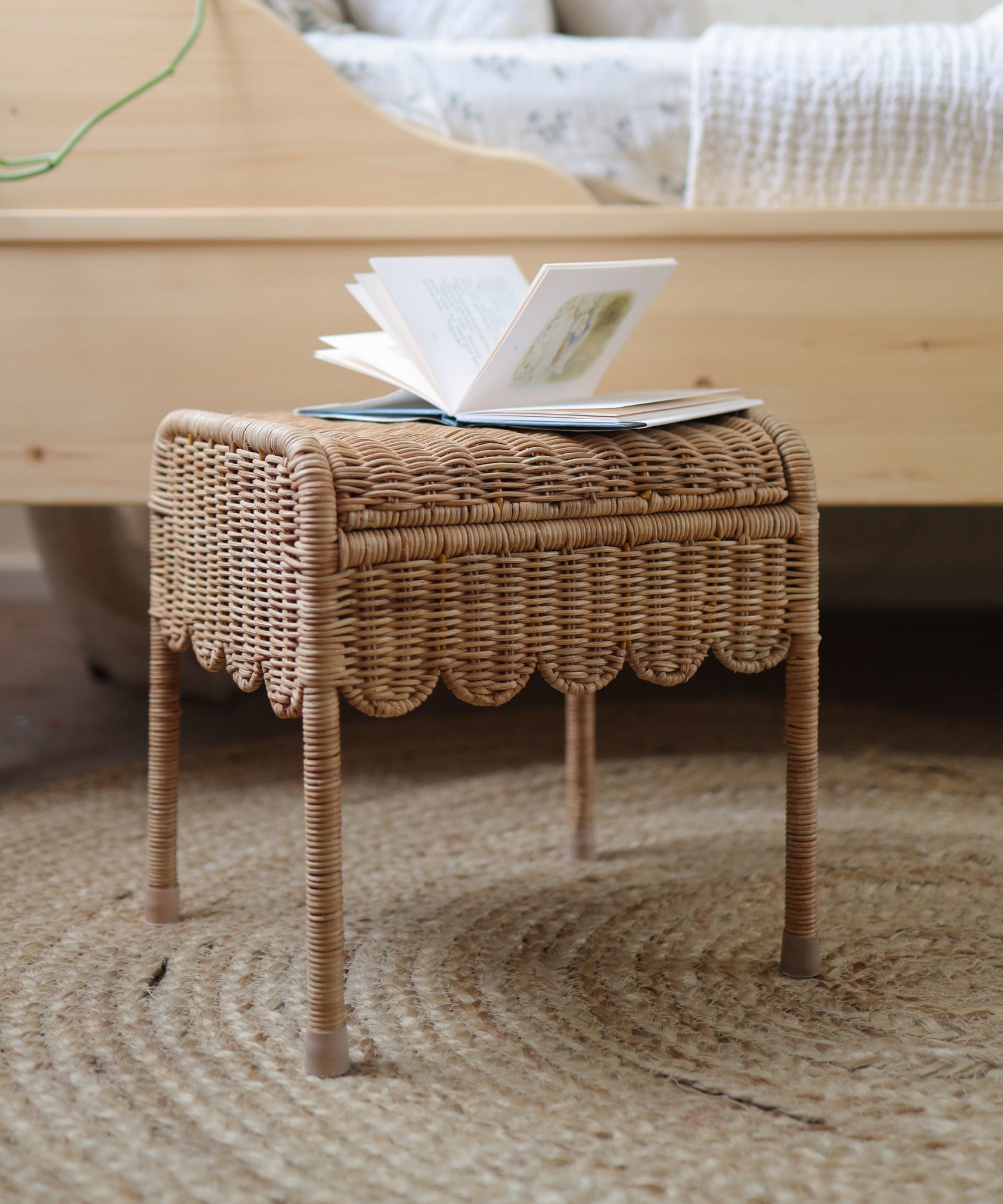 A Olli Ella Petal Storie Stool in natural Rattan. The stool has been placed by the side of a bed with an open book placed on top. 