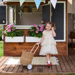 A child standing outdoors in a party dress holding a Natural Olli Ella Rattan Petal Luggy by their side.