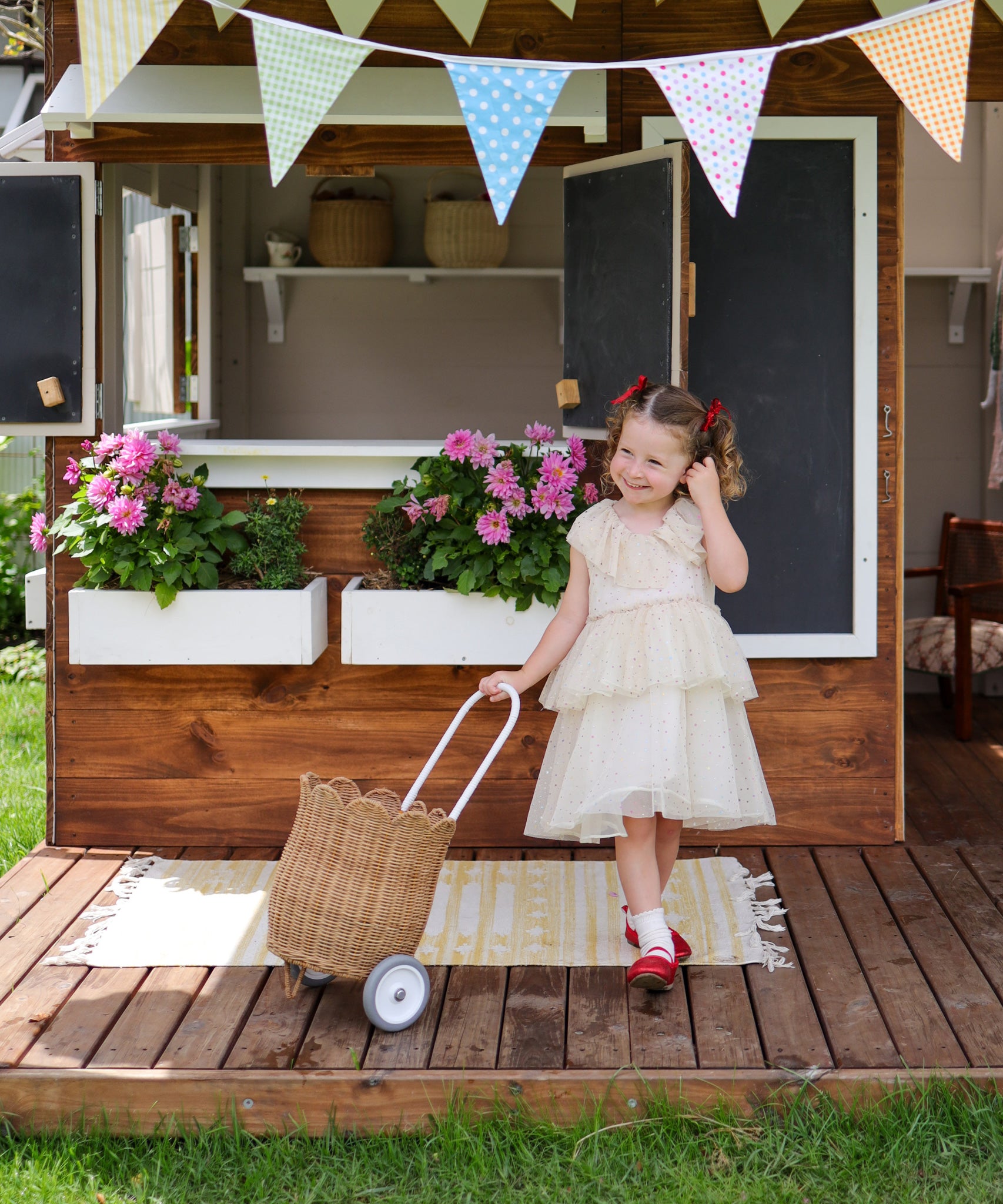 A child standing outdoors in a party dress holding a Natural Olli Ella Rattan Petal Luggy by their side.