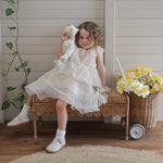 A child sitting on a Natural Rattan Olli Ella Storie Bench. The child is holding a Dozy Dinkum Doll and there is a daffodil filled petal luggy besides the bench.